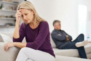 Divorce Lawyer Broward County - Dixon Law Firm Worried woman in a purple top sits on a sofa with her hand on her forehead, while a distant man lounges in the background.