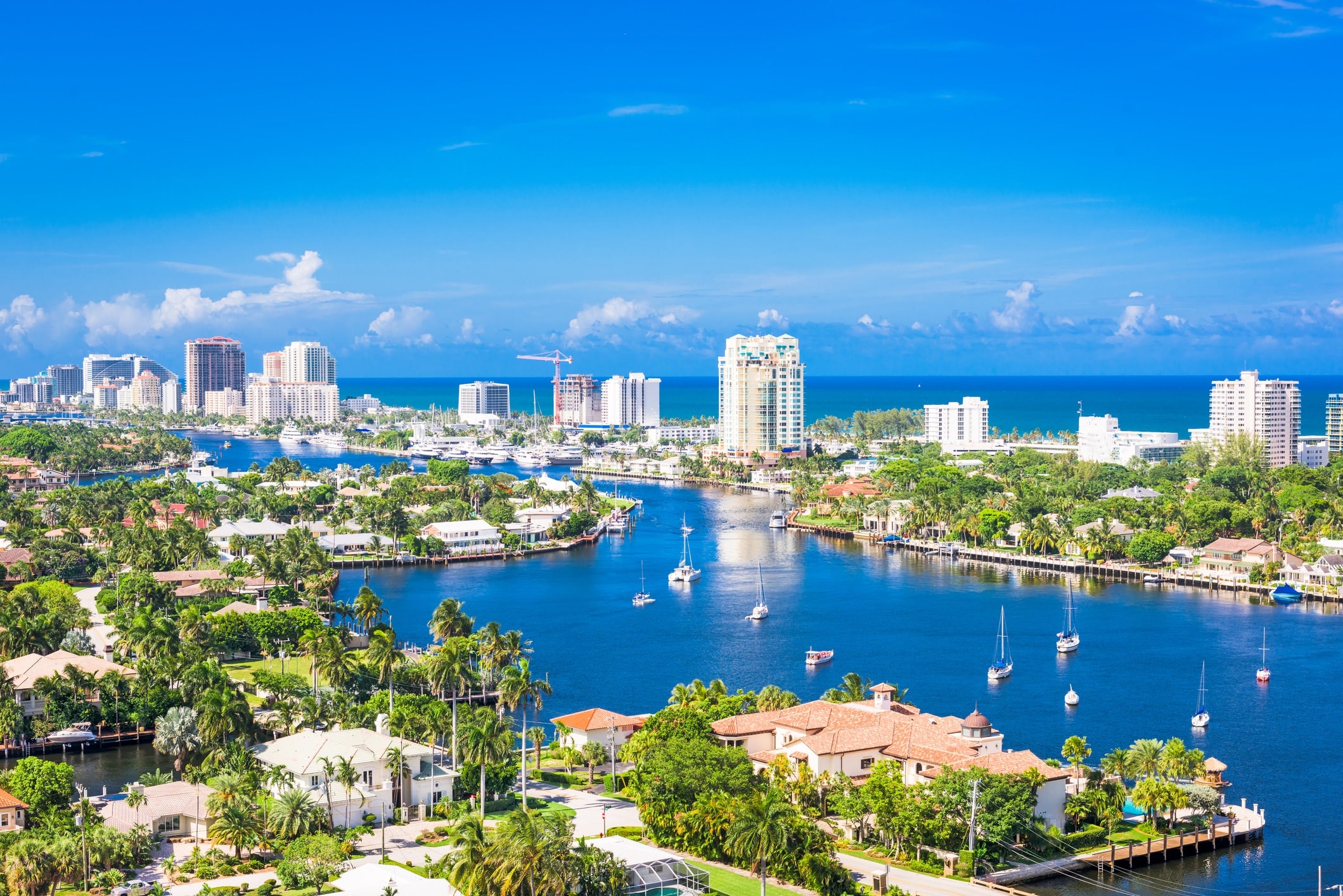 Fort-Lauderdale-South-Florida - Dixon Law Firm A coastal city skyline with high-rise buildings along a marina and turquoise bay, palm trees lining the waterfront and boats in the waterway.