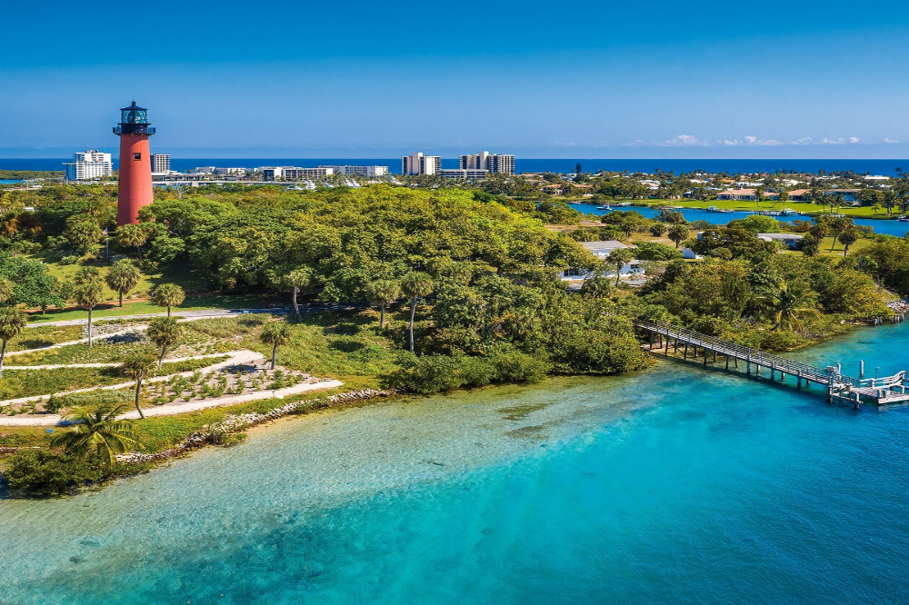 Jupiter - Dixon Law Firm A red lighthouse towering over lush green coastal landscape with a wooden pier extending into turquoise water and a distant city skyline on the horizon.