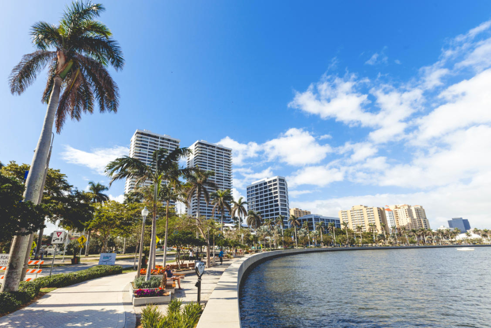 West Palm Beach - Dixon Law Firm Palm-lined waterfront promenade with modern high-rise buildings along the water under a bright blue sky, palm trees swaying in the breeze.