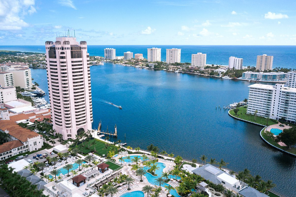 Boca Raton FL - Dixon Law Firm Aerial view of a coastal resort with a pink high-rise, blue water canal, and white apartment buildings along the shoreline, under a bright sky