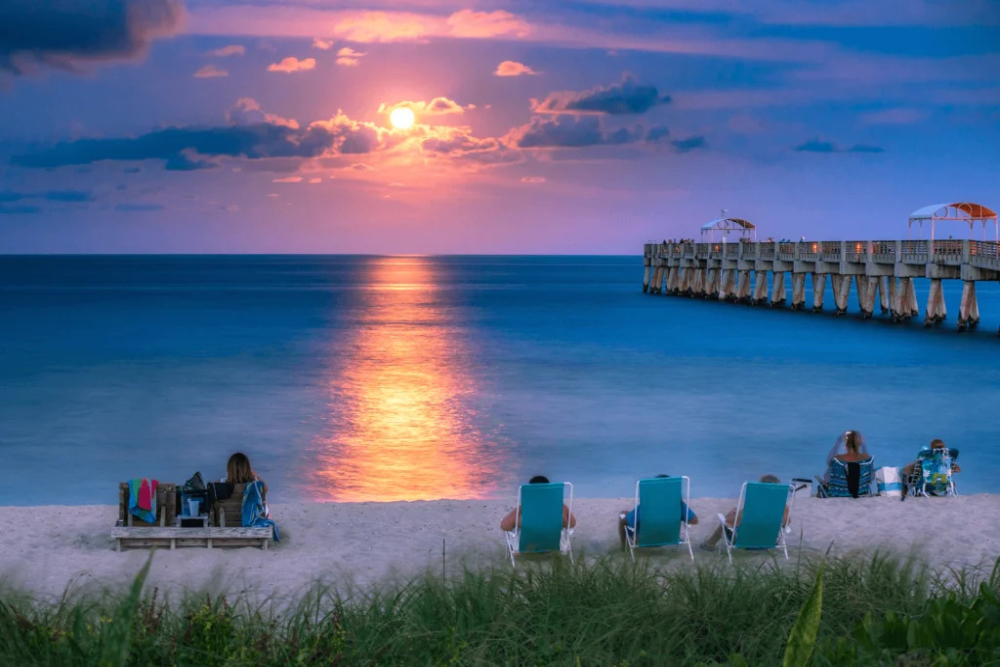 Lake Worth - Dixon Law Firm Sunset over the ocean with a long pier jutting into the water; beachgoers relax on chairs and blankets on the sand.