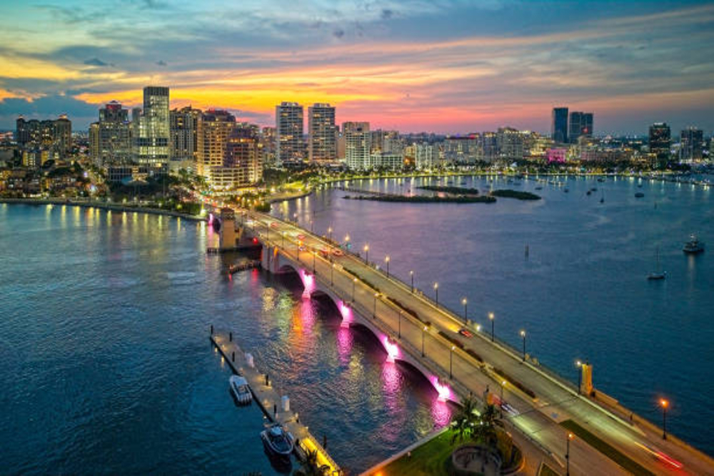 Palm Beach - Dixon Law Firm City skyline at sunset with a lit bridge extending over calm water, reflecting pink lights from the arches.