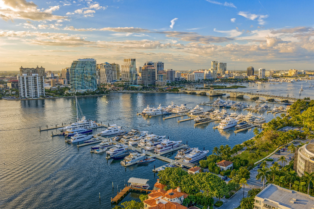Royal Palm Beach - Dixon Law Firm Aerial view of a marina with many yachts docked at piers, calm water, and a modern city skyline across the bay under a blue sky.